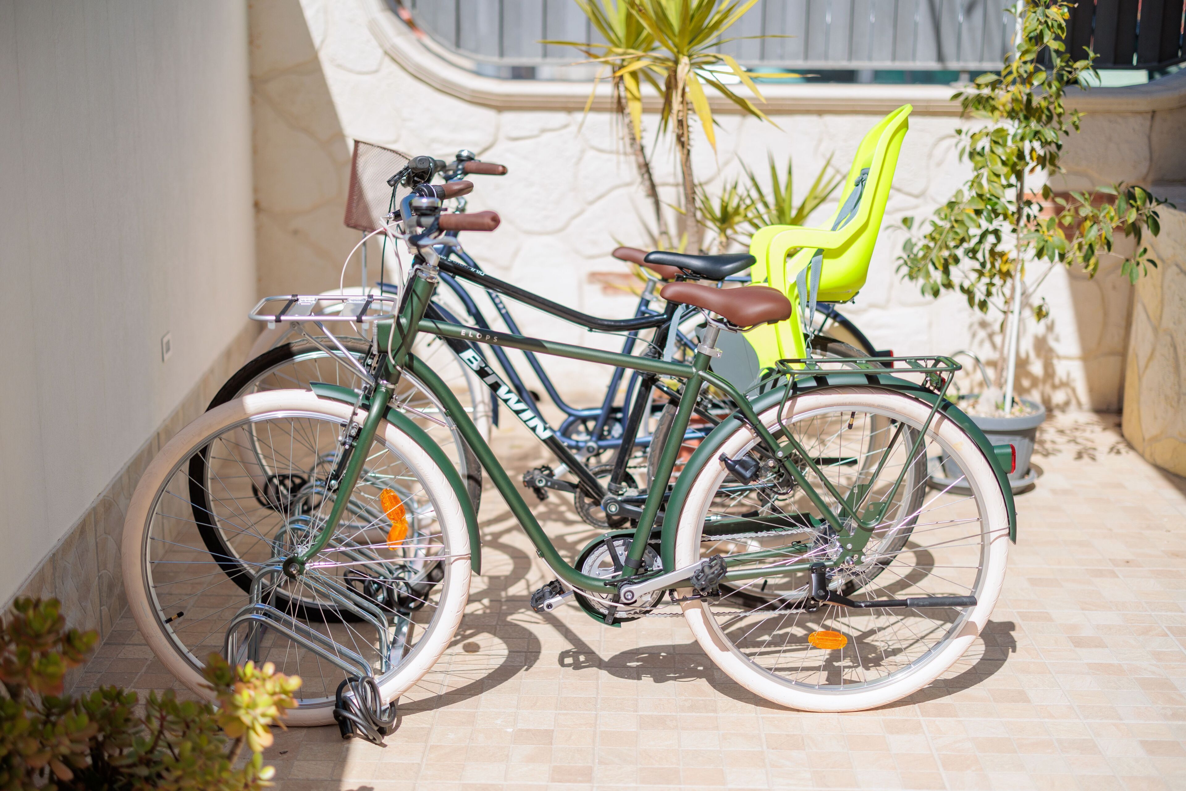 A bicycle parked next to a chair and a potted plant in a sunlit outdoor area with a tiled floor.