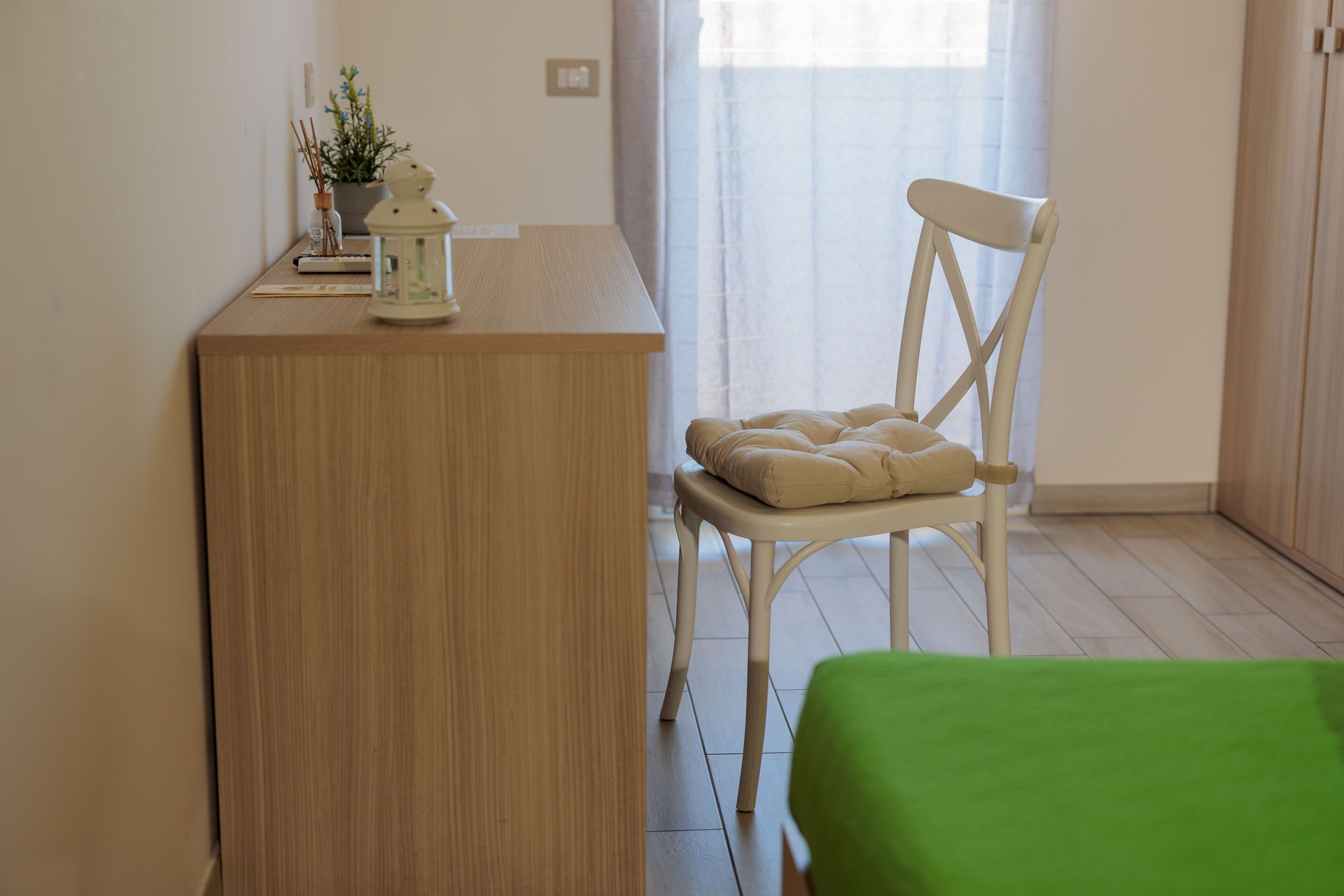 Una habitación con un escritorio de madera, una silla blanca con cojín, una cama verde, una planta en maceta y una ventana con cortinas traslúcidas.