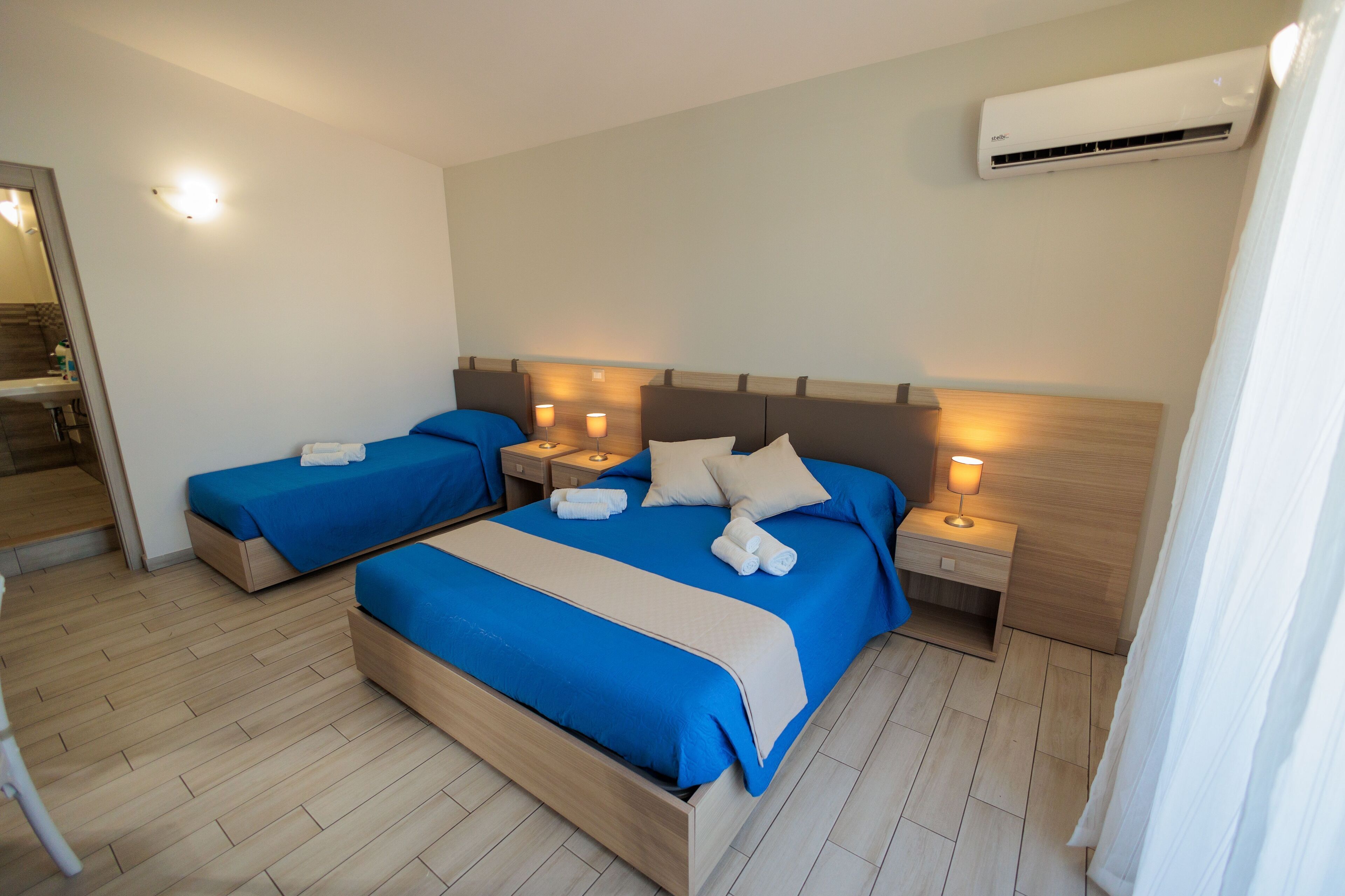 A hotel room with two beds, each with blue bedding and white towels, a wooden headboard, bedside lamps, and an air conditioning unit on the wall.
