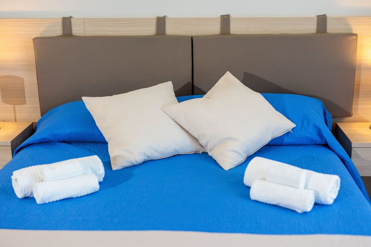 A bed with a blue bedspread, two white pillows, and two rolled white towels.