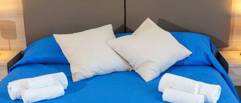 Una cama con una colcha azul, dos almohadas blancas y dos toallas blancas enrolladas.
