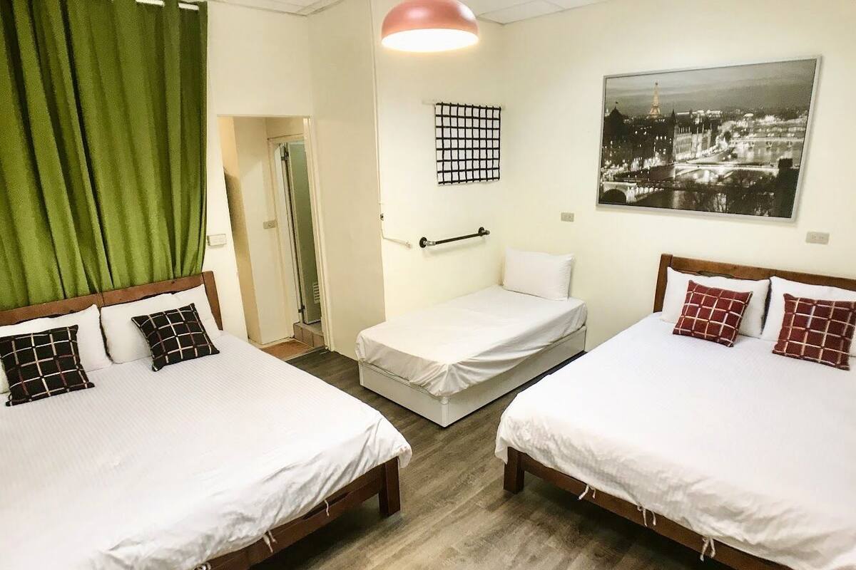 A hotel room with two single beds, a small bedside table, a green curtain, a framed cityscape picture, and a grid-patterned wall decoration.