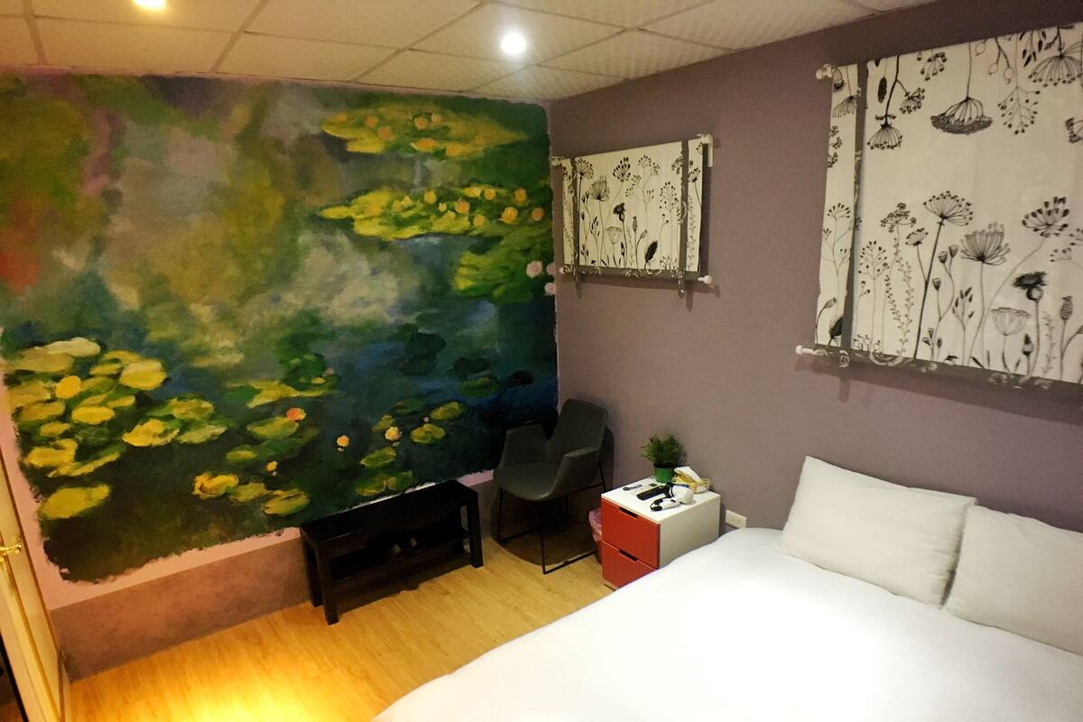 A bedroom with a large mural of a pond and flowers on the wall, a bed, a desk with a chair, and a small table with a plant.