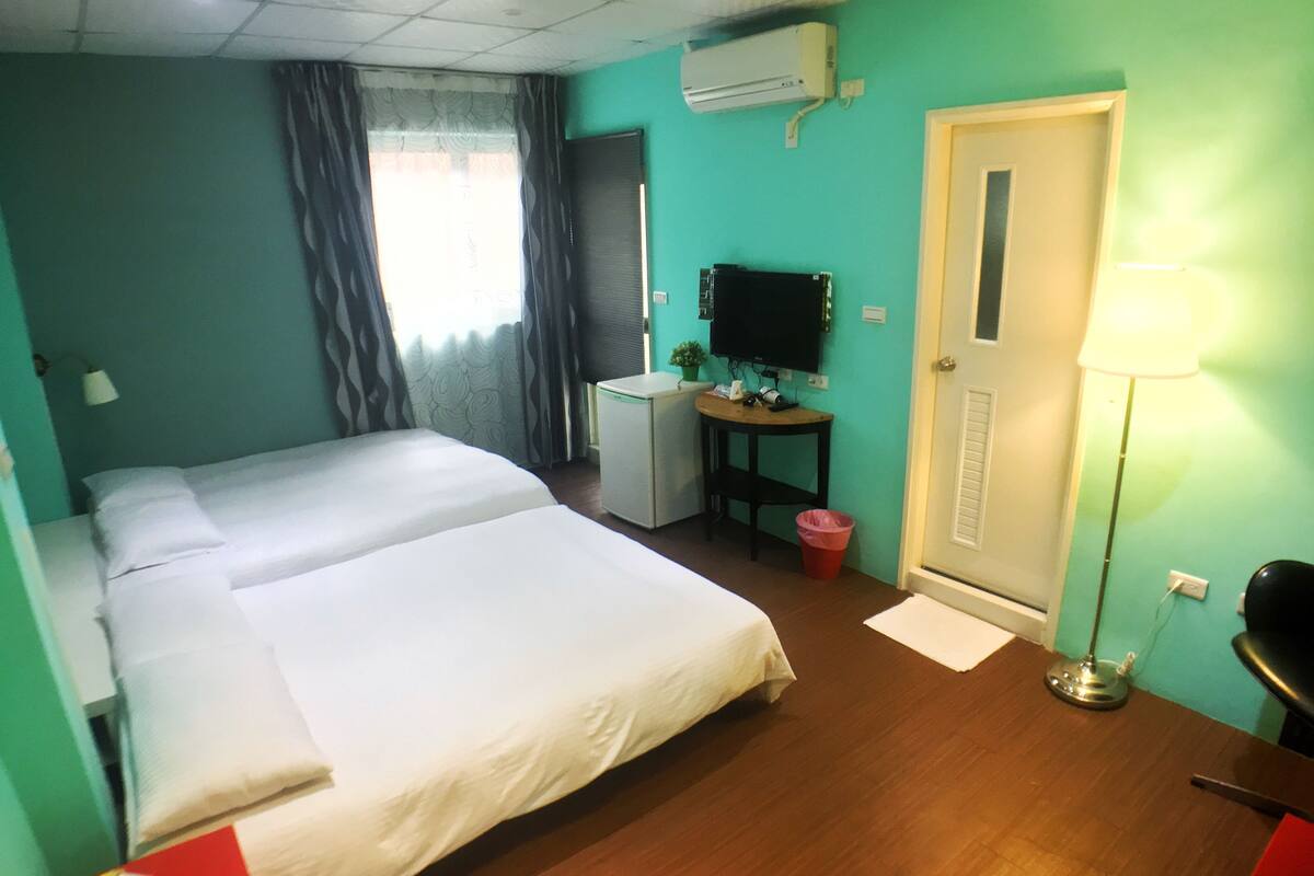 A hotel room with two beds, a desk with a computer, an air conditioner, and a door.