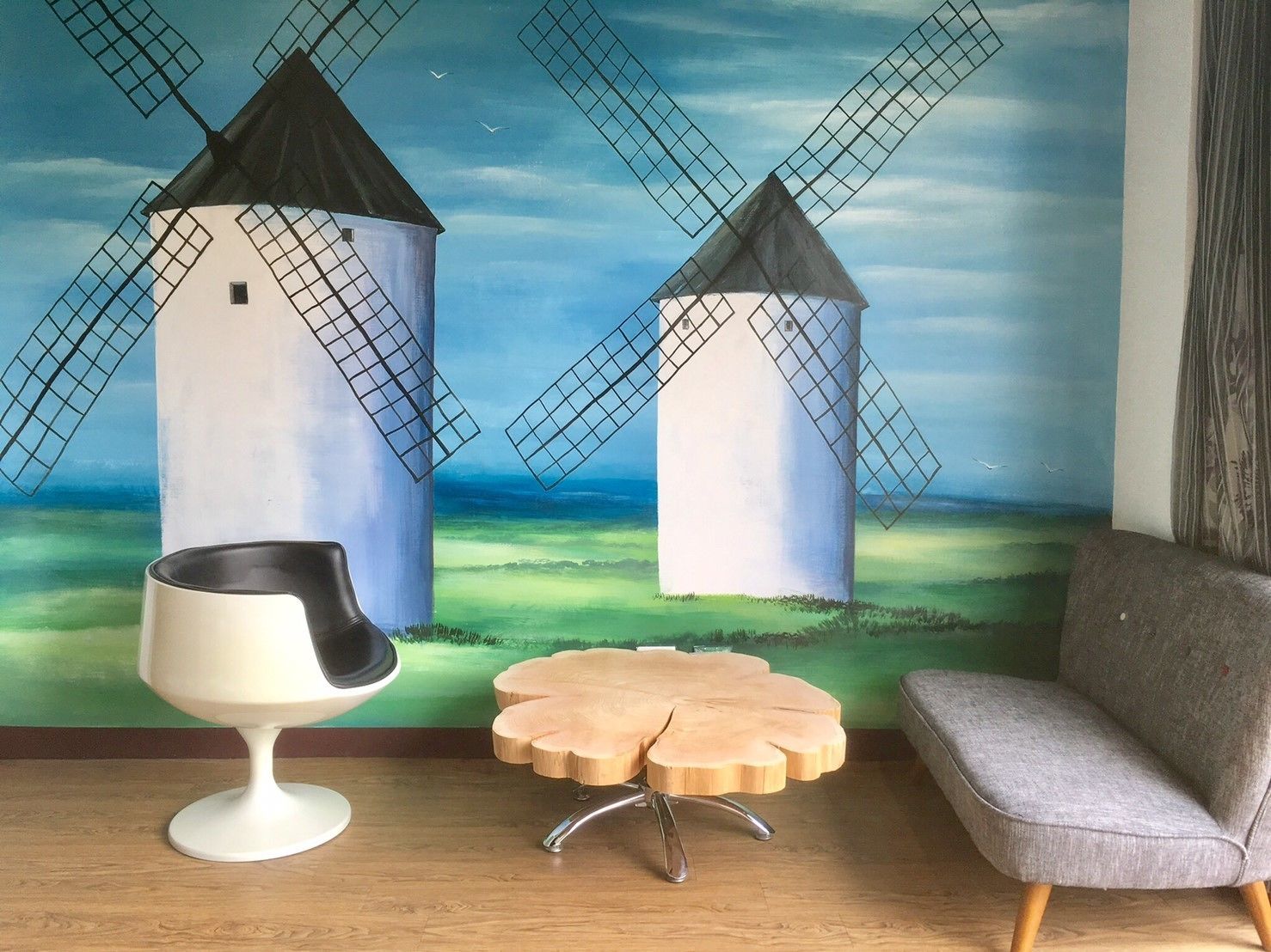 A room with a mural of windmills, a white chair, a wooden table, and a grey sofa.