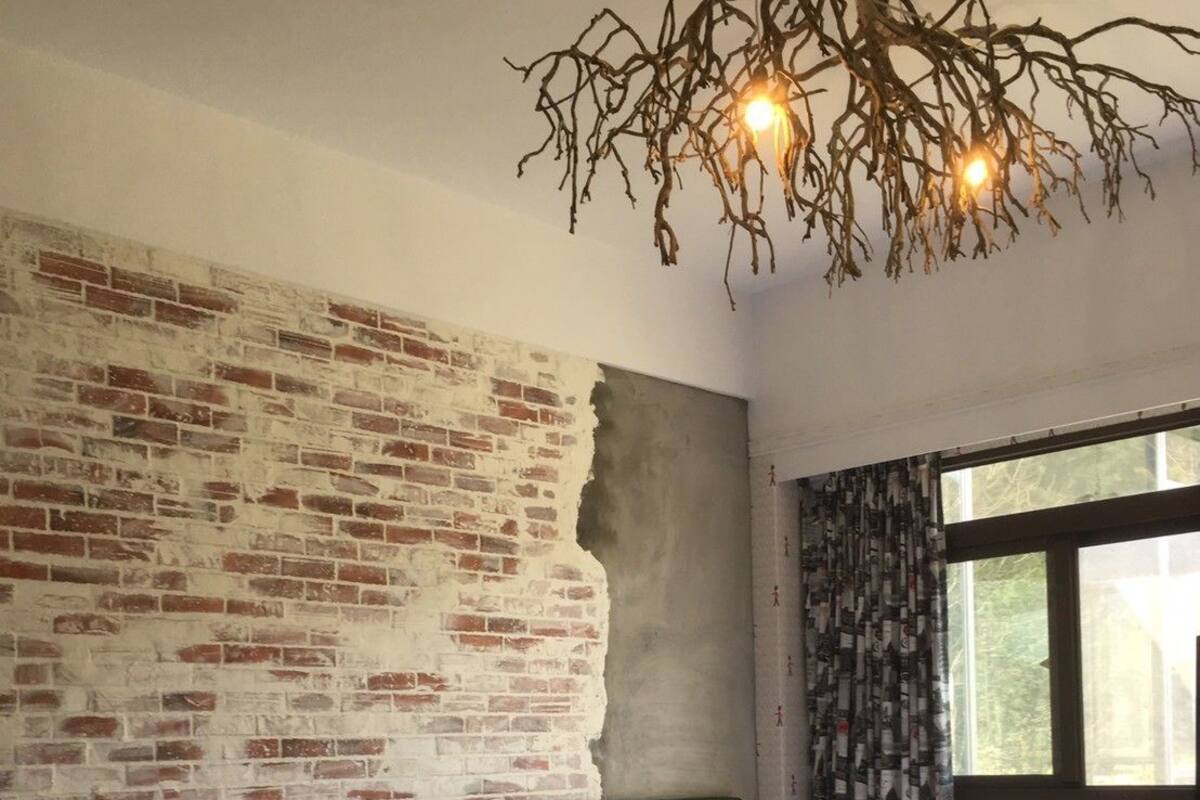 Una habitación con dos camas, una pared de ladrillo, un cabecero verde, una ventana con cortinas y una lámpara colgante.