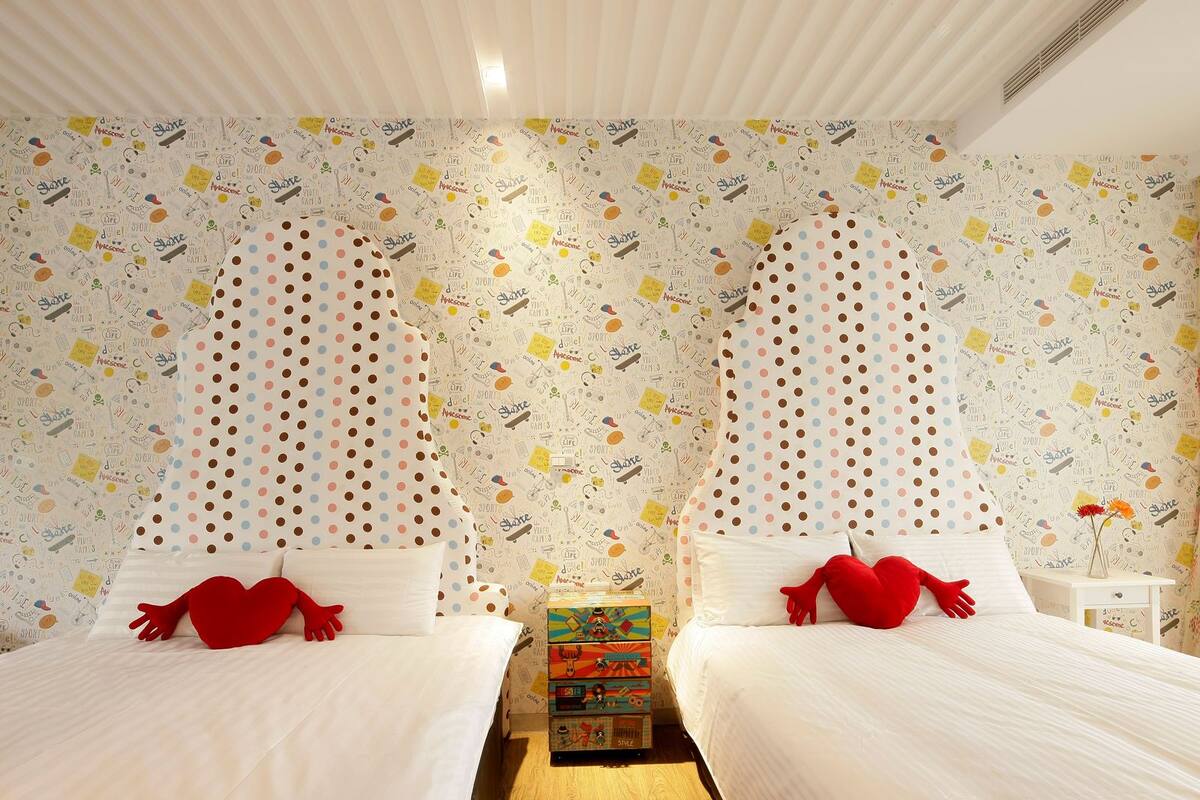 Dormitorio con dos camas, cada una con una almohada en forma de corazón rojo, y papel tapiz estampado con garabatos y palabras.