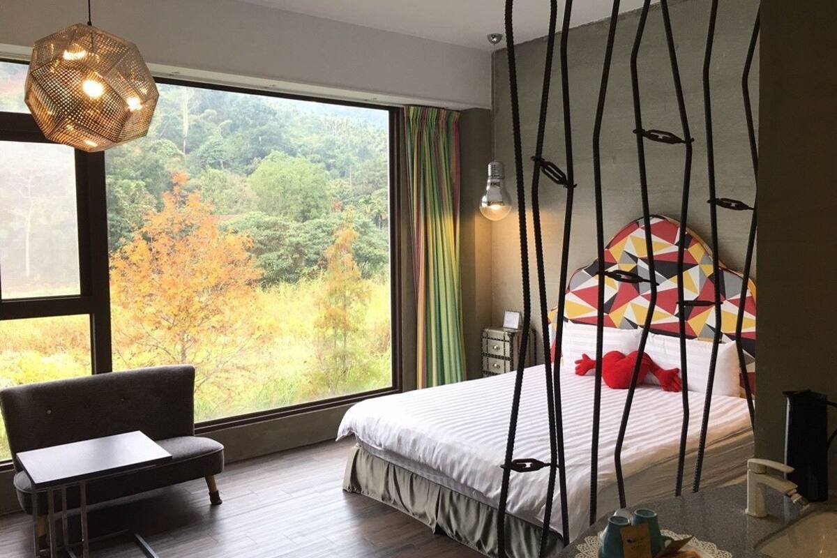 Un dormitorio con un ventanal grande, una cama con cabecera geométrica colorida, una lámpara colgante y una mesita.