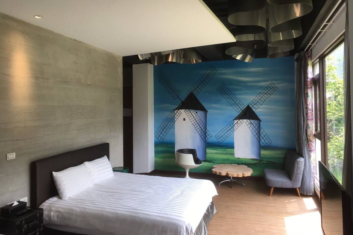 Una habitación de hotel moderna con una cama grande, un mural de molinos y una mesa redonda con una silla.