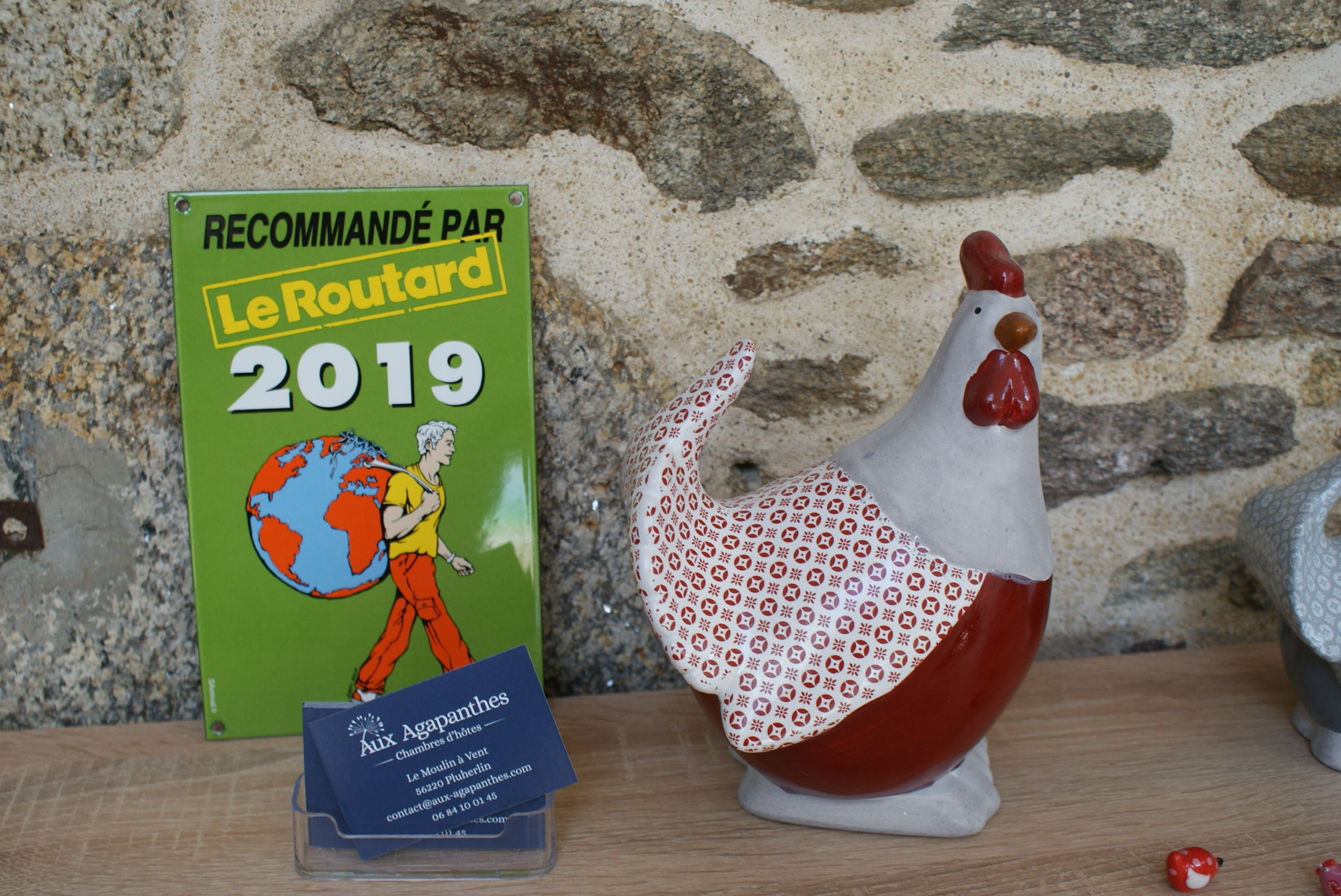 Una figurita de cerámica de gallo y un libro titulado "RECOMMANDÉ PAR Le Routard 2019" sobre una mesa frente a una pared de piedra.