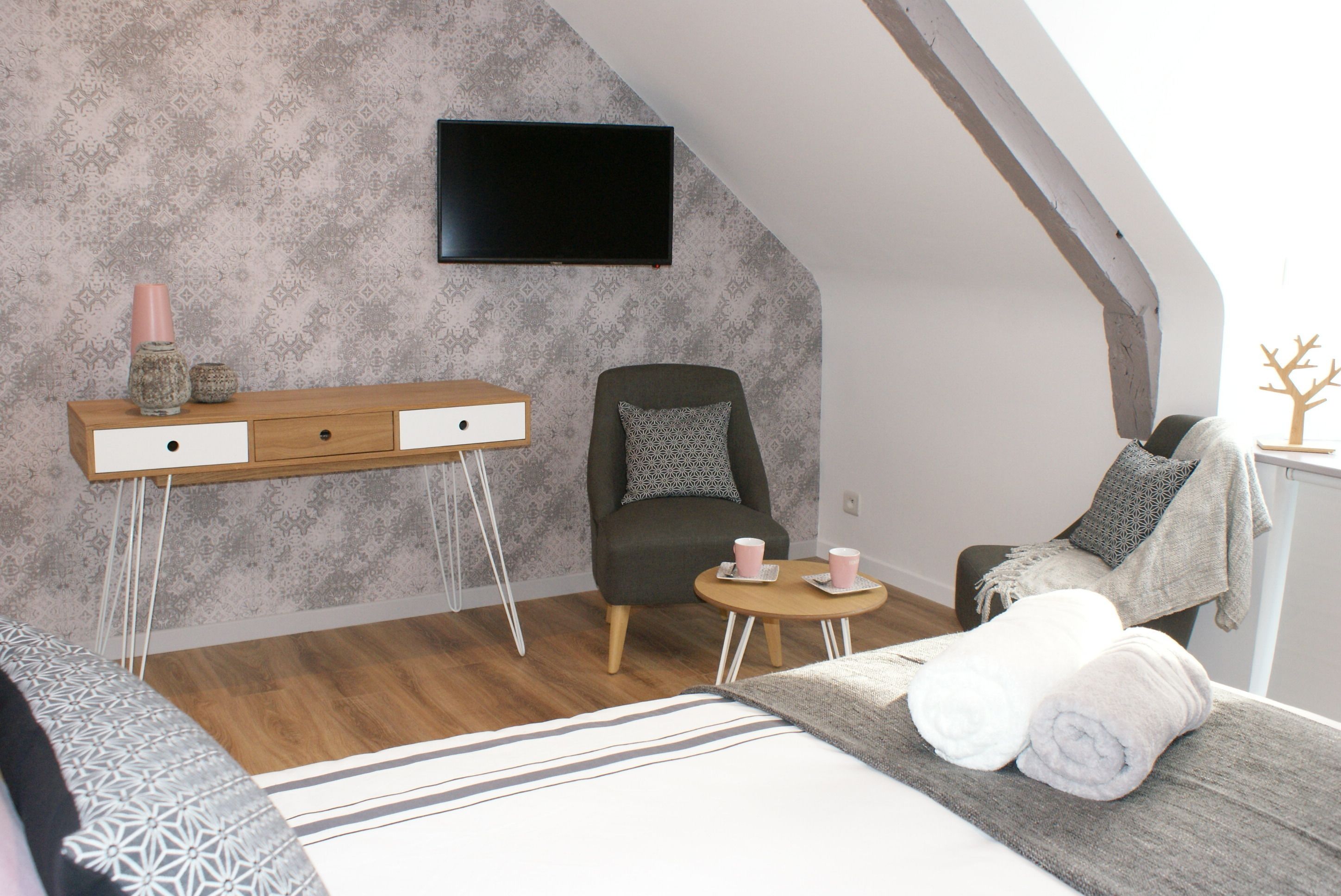 A modern bedroom with a loft-style ceiling, a bed with a striped bedspread, a wooden bedside table with a pink vase, a gray armchair, and a small round table with two pink cups.