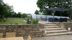 A patio area with a stone wall, wooden table and chairs, a glass greenhouse structure, and outdoor seating with a pergola. - Aux Agapanthes (Pluherlin)