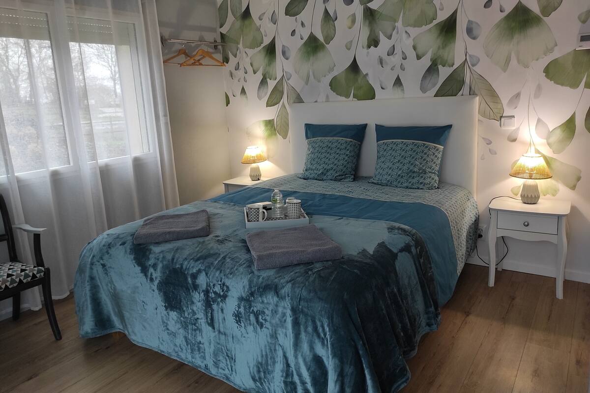 Un dormitorio con una cama grande, dos mesitas de noche, una silla y un ventanal con cortinas translúcidas.