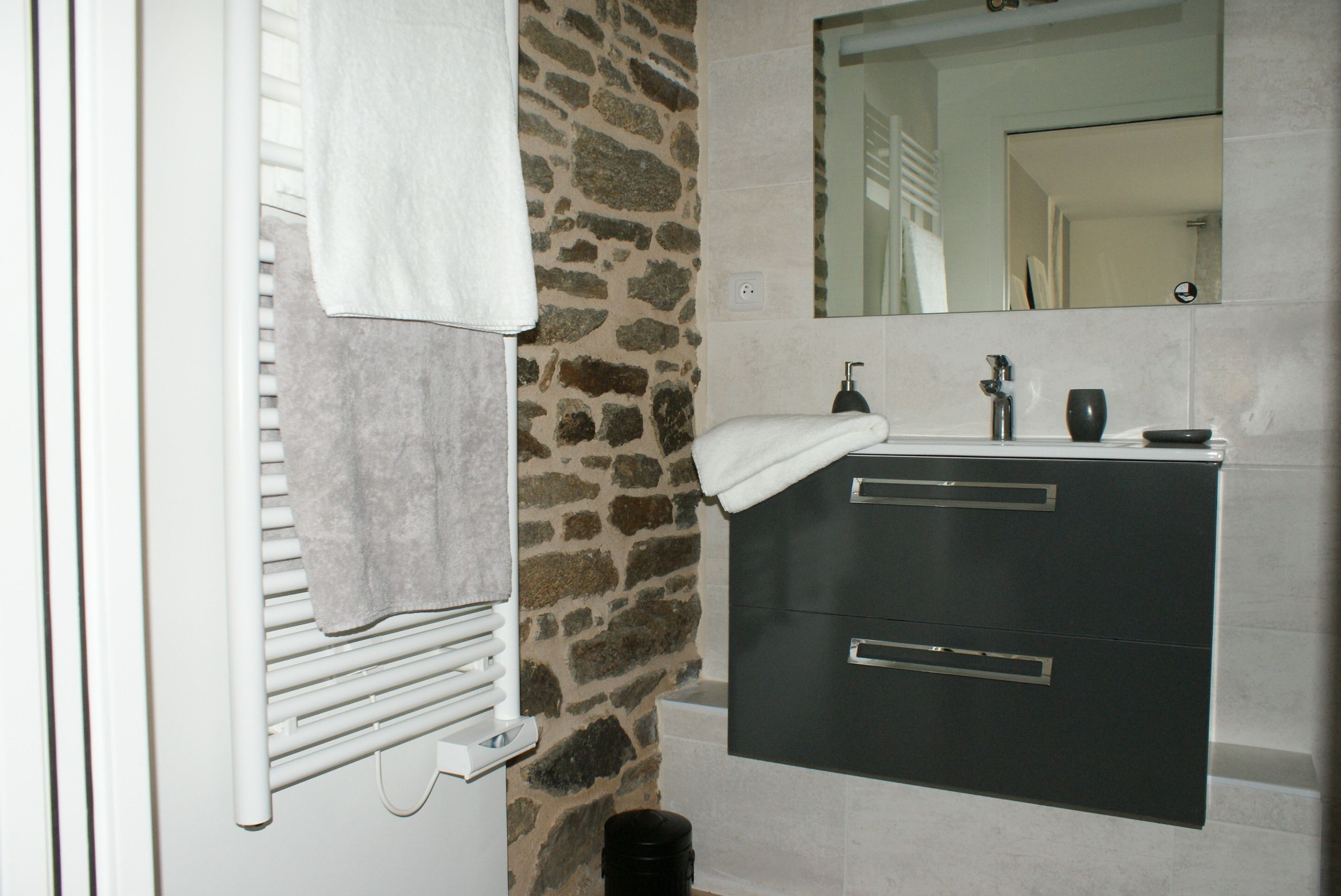 A bathroom with a stone wall, a white towel hanging on a radiator, a black sink with a mirror above it, and a black trash bin.