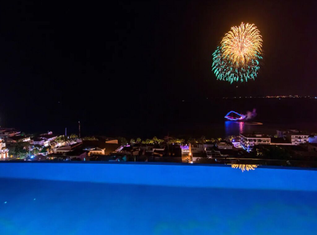 Une vue nocturne d’une ville côtière, illuminée par un grand spectacle de feux d’artifice dans le ciel.