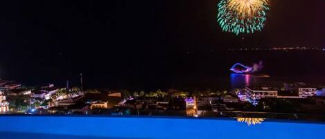 Une vue nocturne d’une ville côtière, illuminée par un grand spectacle de feux d’artifice dans le ciel.