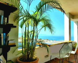 A balcony with a view of the sea, a potted plant, a metal chair, and a bookshelf with books. - OCEAN VIEWS~2Beds~Steps to Beach & Malecon~Close to Everything ~Safe Loc (5 de Diciembre)