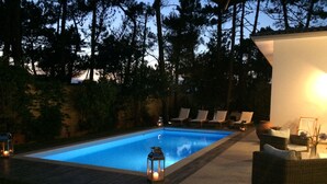 Una piscina rectangular con borde de baldosas, rodeada de sillas de descanso y una pérgola con una lámpara.