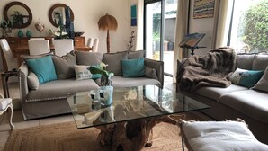 Una sala de estar moderna con una mesa de centro de vidrio, dos sofás con cojines color turquesa, una mesita de noche de madera y un ventanal con vistas a la vegetación.