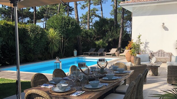 Un área de comedor al aire libre con mobiliario de mimbre, una piscina y un edificio blanco al fondo.