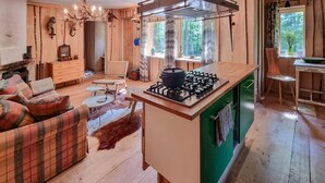 A cozy cabin interior with a fireplace, a dining area, a kitchen with a gas stove, and a large window overlooking a forest.