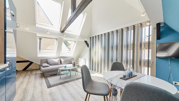 A modern living room with a sofa, coffee table, and dining table. There are large windows and a TV mounted on the wall. - Apartments am Markt (Bernkastel-Kues)