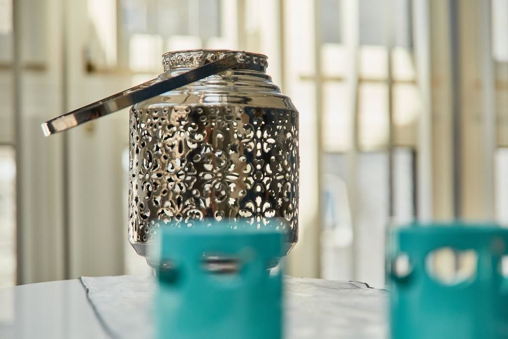 Un récipient métallique orné de motifs complexes et doté d’une anse, posé sur une surface où se trouvent des chaises de couleur turquoise.