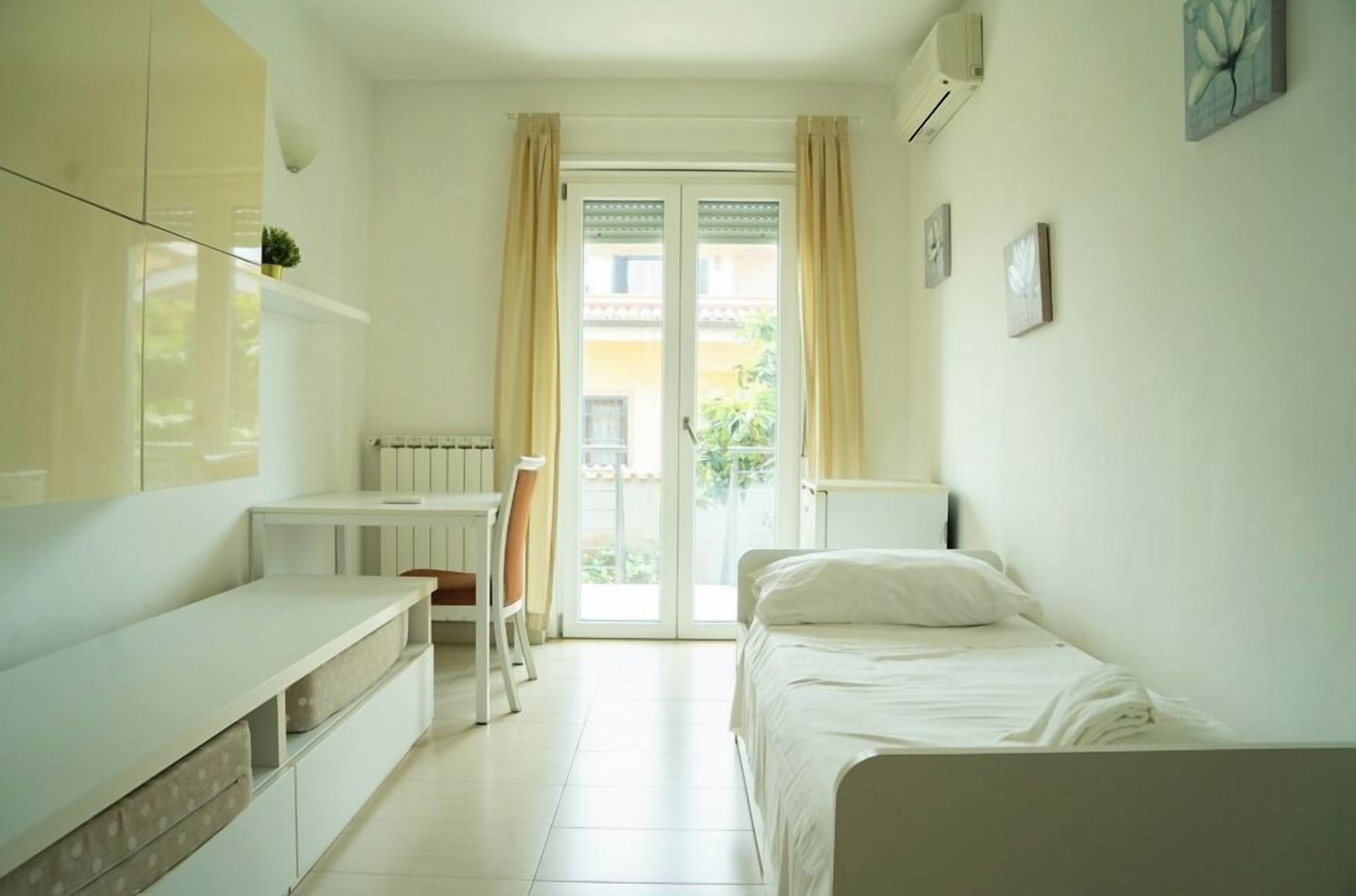 A small, bright room with a bed, a desk, and a chair. There is a window with curtains and a radiator on the wall.