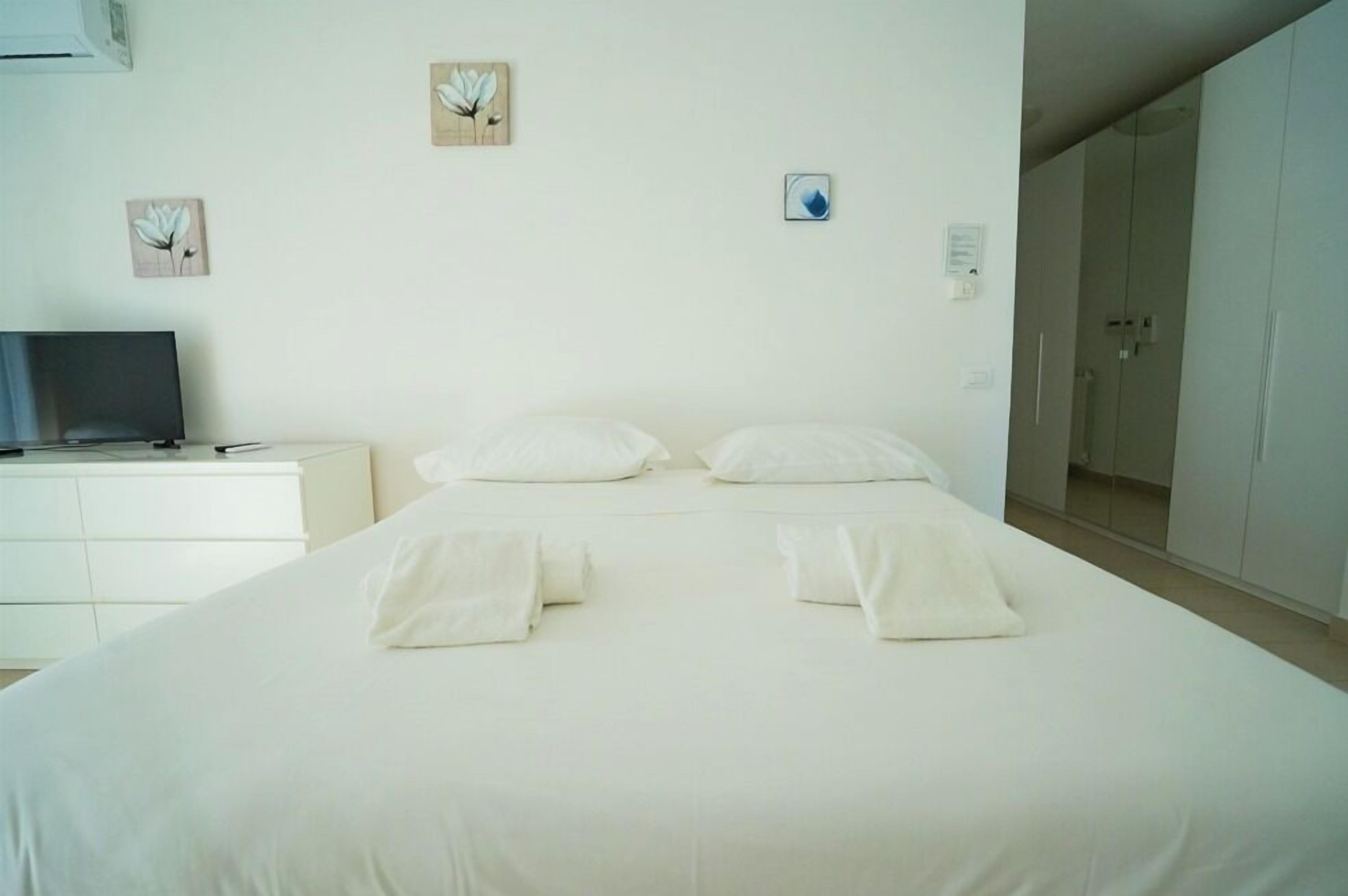 A neatly made bed with pillows and folded towels in a clean, white room with a television on a white dresser.