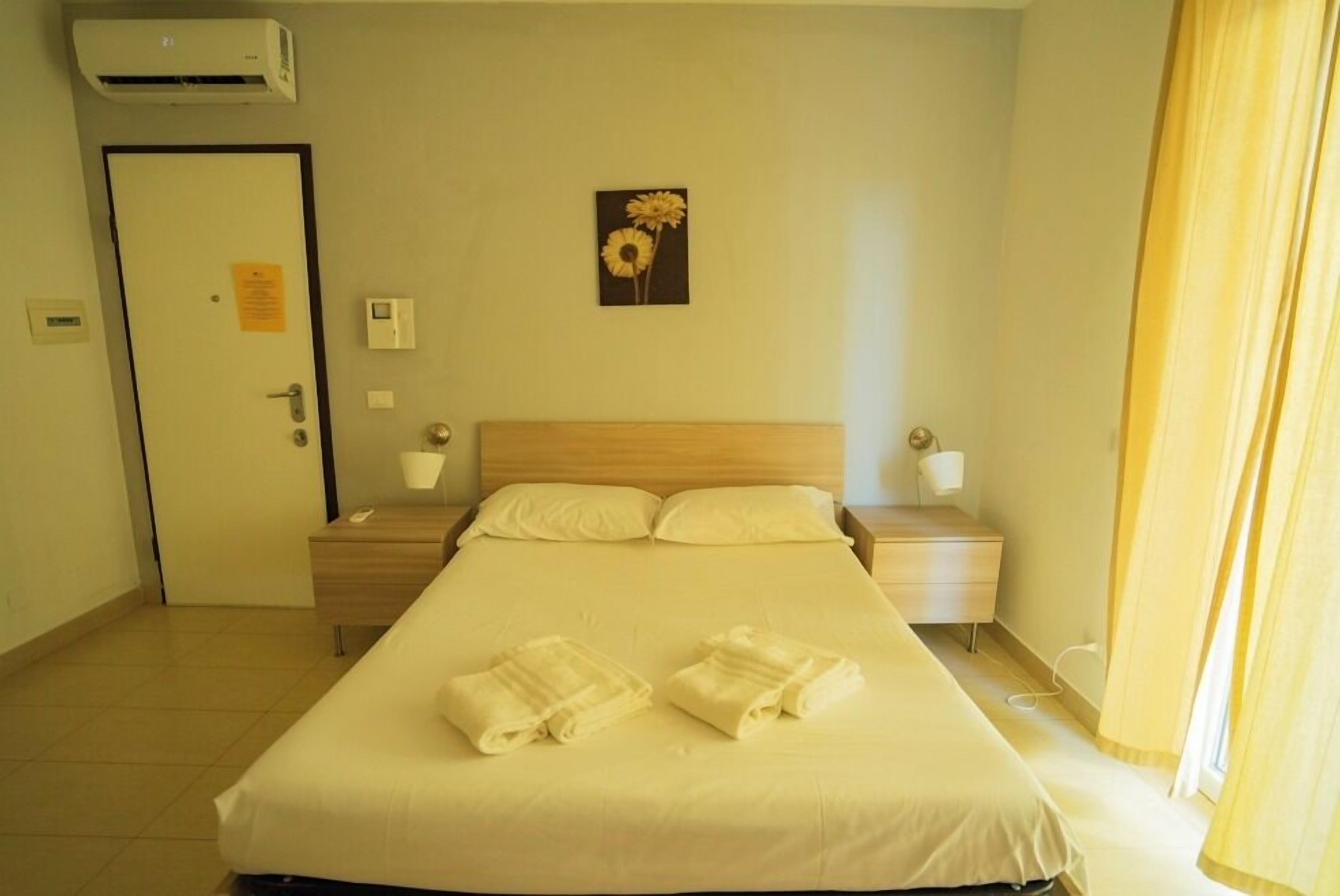 Een hotelkamer met een bed, twee nachtkastjes, een airconditioning, een deur, een lichtschakelaar aan de muur en een schilderij van bloemen.