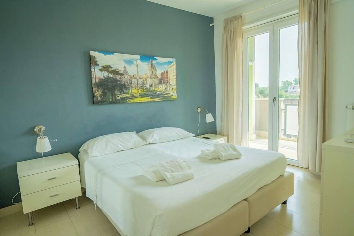 A hotel room with a bed, a nightstand, a painting on the wall, and a view of the outside.