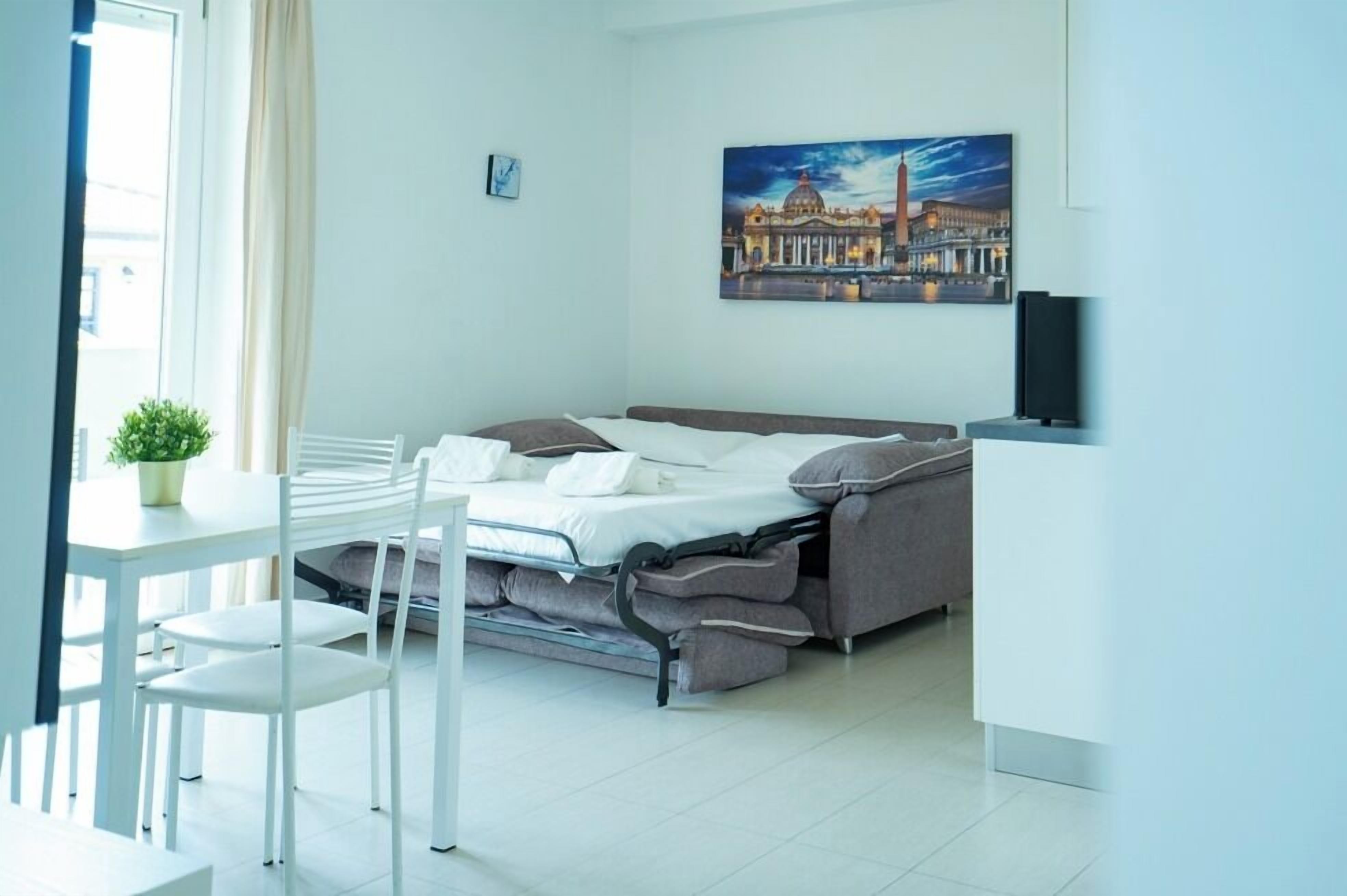 A compact hotel room with a bed, a small table with chairs, a potted plant, and a painting of a building on the wall.