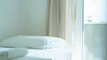 A neatly made bed with white linens in a room with a large window and sheer curtains.