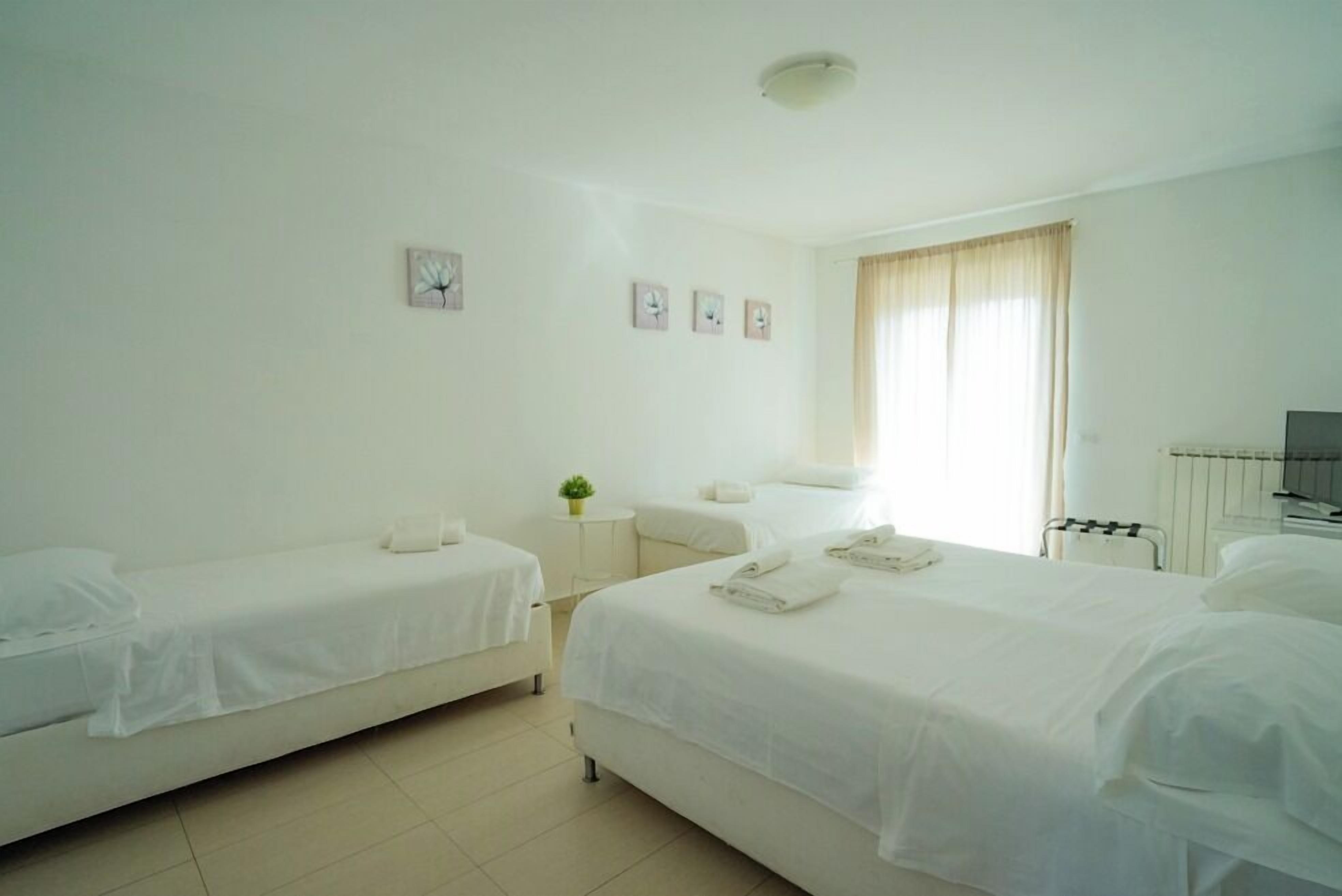 A hotel room with two single beds, a small bedside table with a lamp, a large bed with a pillow and a blanket, a window with curtains, and a wall clock.