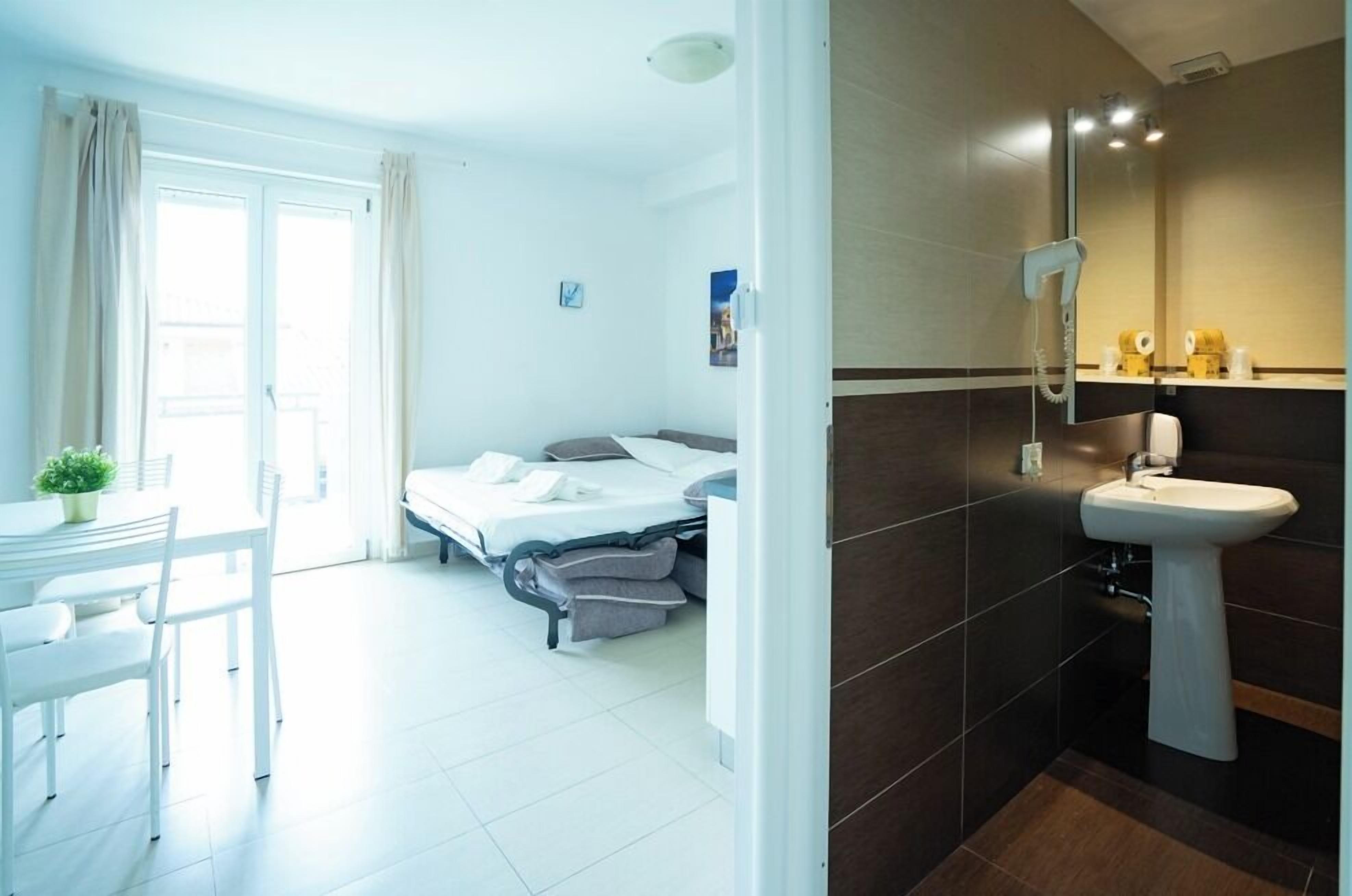A hotel room with a bed, a desk with a chair, a small table with a plant, a bathroom with a sink and mirror, and a shower.