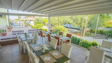 Un espacio cubierto para comer al aire libre con varias mesas y sillas, plantas en macetas y vistas a la calle y al verdor.