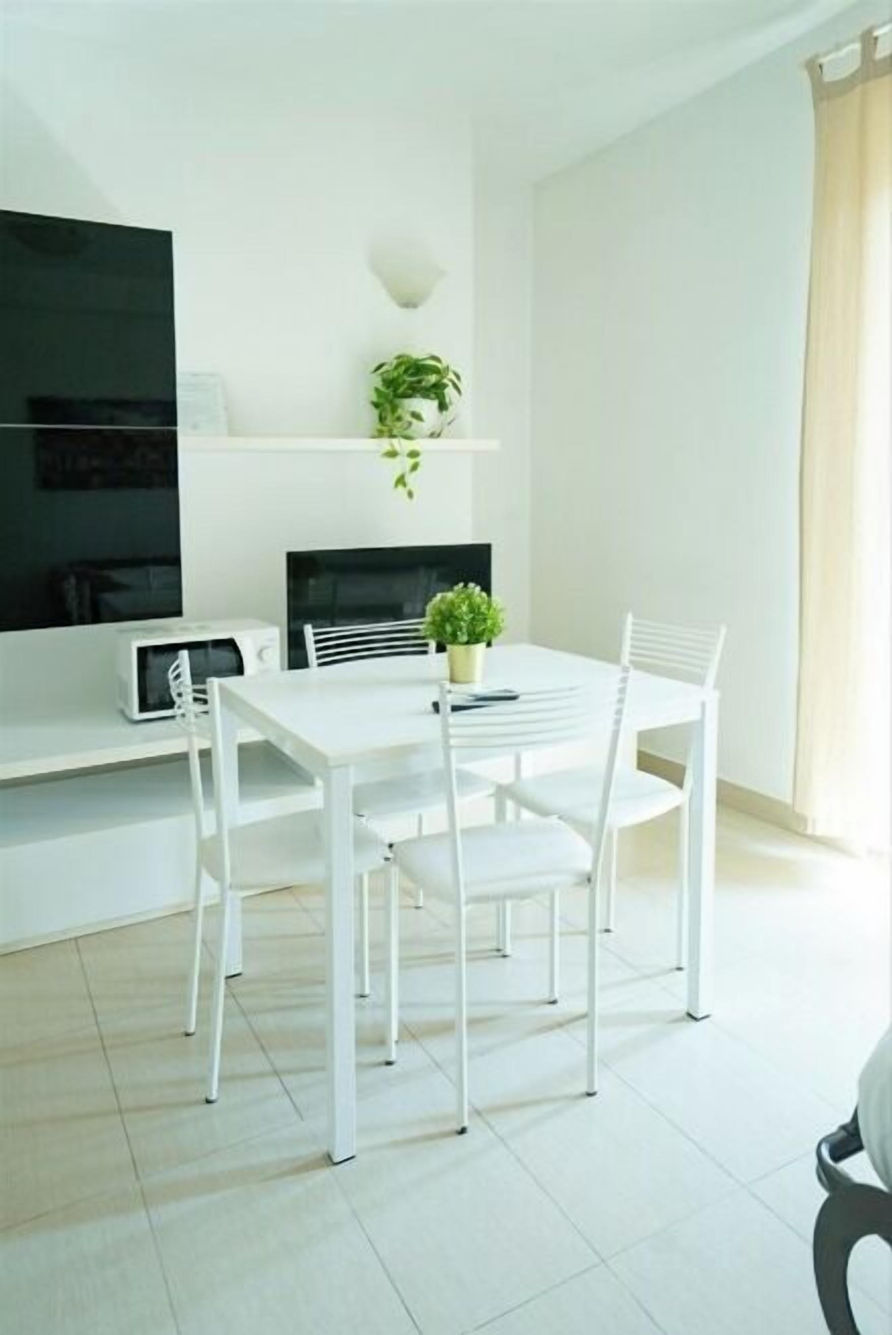 A modern, minimalist dining area with a white table and chairs, a small potted plant, and a flat-screen TV on a white stand.