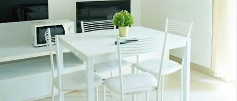 A modern, minimalist dining area with a white table and chairs, a small potted plant, and a flat-screen TV on a white stand.