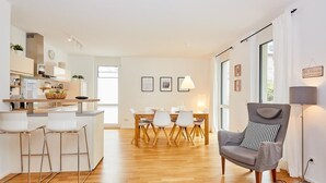 A modern kitchen with white cabinets, a wooden dining table, and bar stools. There is a grey armchair with a patterned cushion in the foreground. - Auszeit (Bernkastel-Kues)