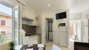 A modern kitchen with white cabinets, a built-in stove, and a microwave. There is a dining area with a table set for two, a potted plant, and a view of the outside.