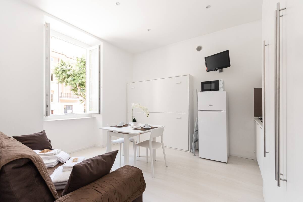 Una camera moderna e luminosa con un letto, un tavolo da pranzo con sedie, un frigorifero bianco e una TV a schermo piatto montata a parete.
