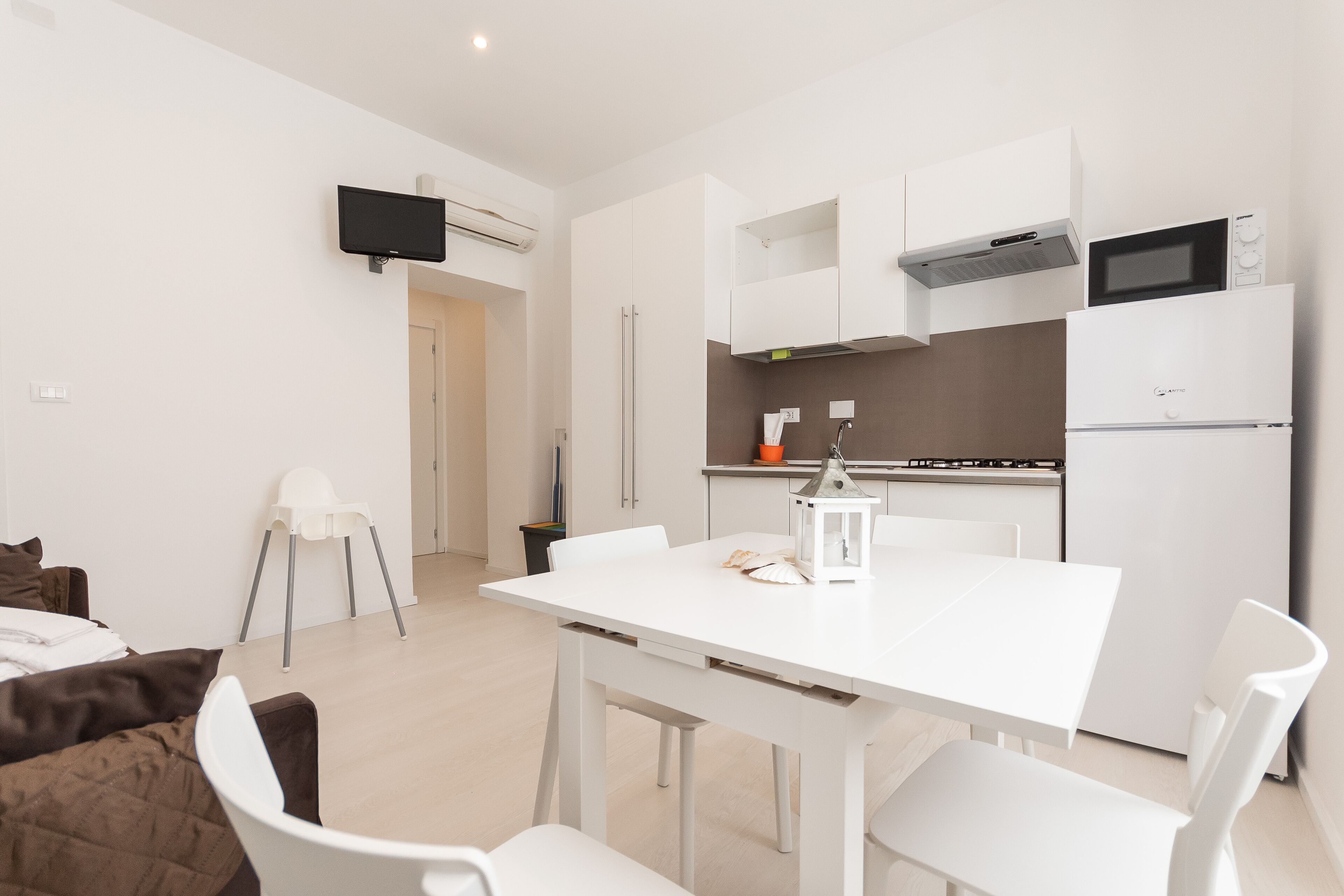 Une cuisine moderne avec des meubles blancs, un four à micro-ondes et un four encastré. On y trouve une table à manger avec des chaises, un téléviseur fixé au mur et une petite chaise blanche dans un coin.
