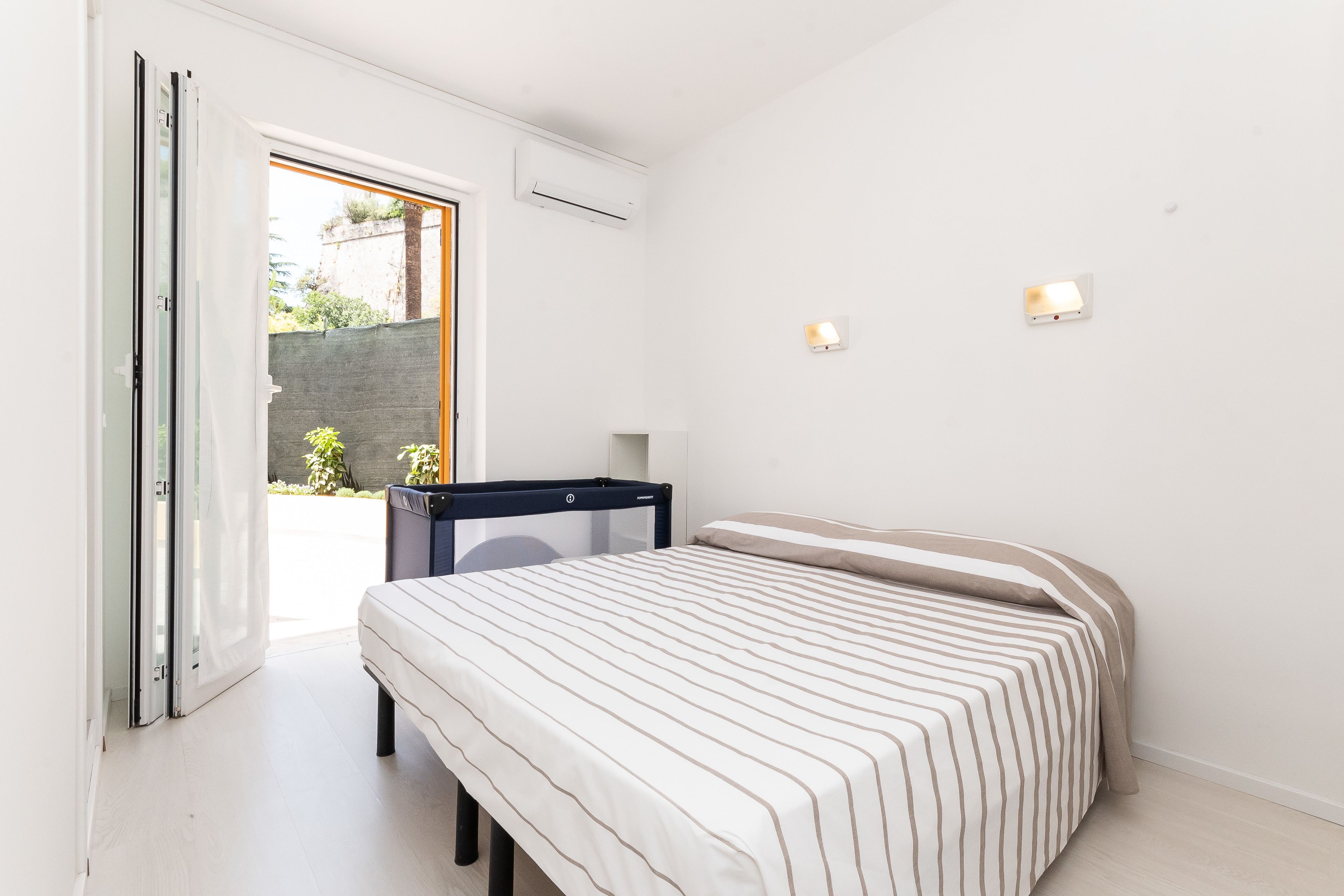A bedroom with a bed, a desk, and a chair. There is a door leading to an outdoor area.
