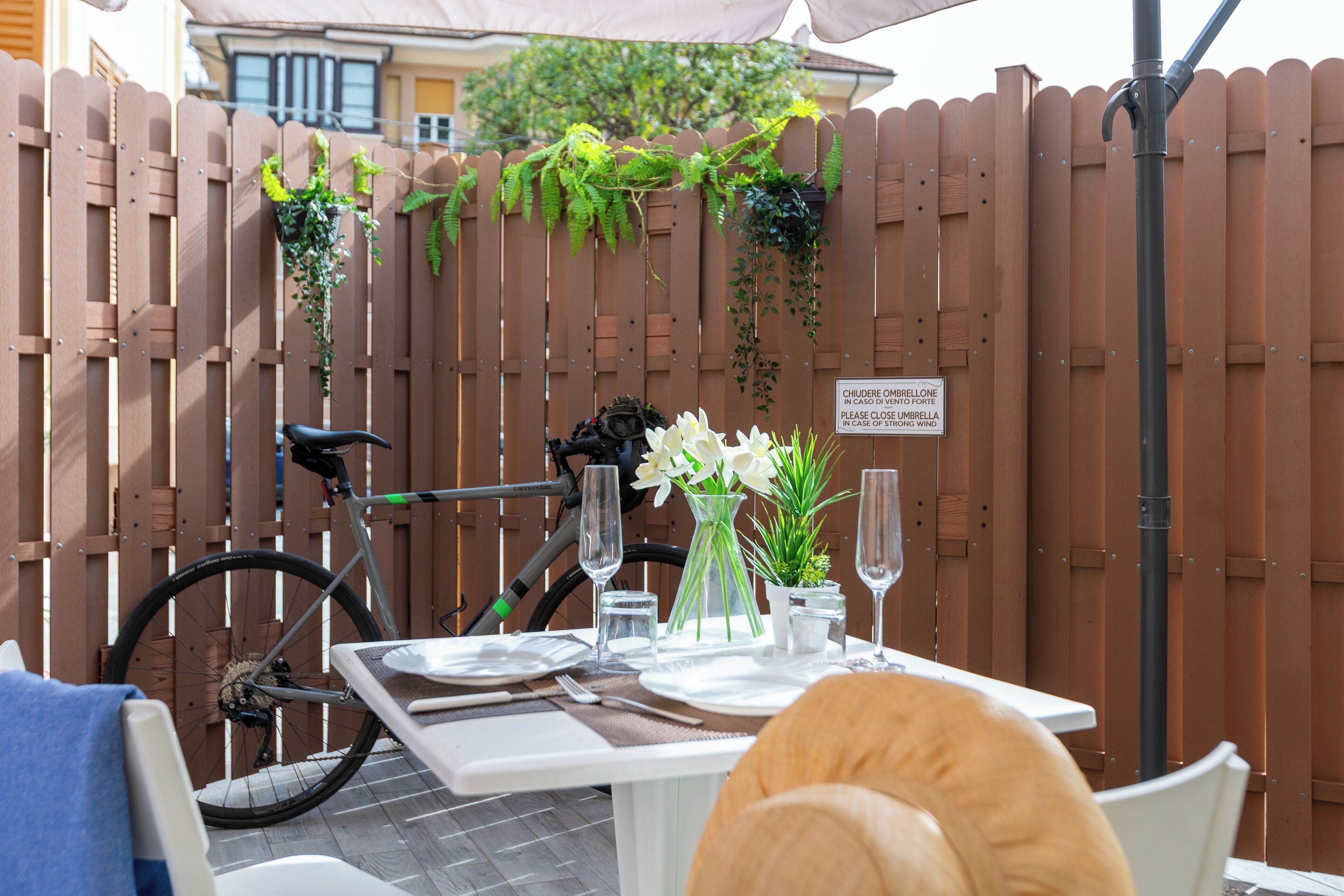 Un'area pranzo all'aperto con recinzione in legno, una bicicletta e un tavolo imbandito per due, con piatti, posate e bicchieri.