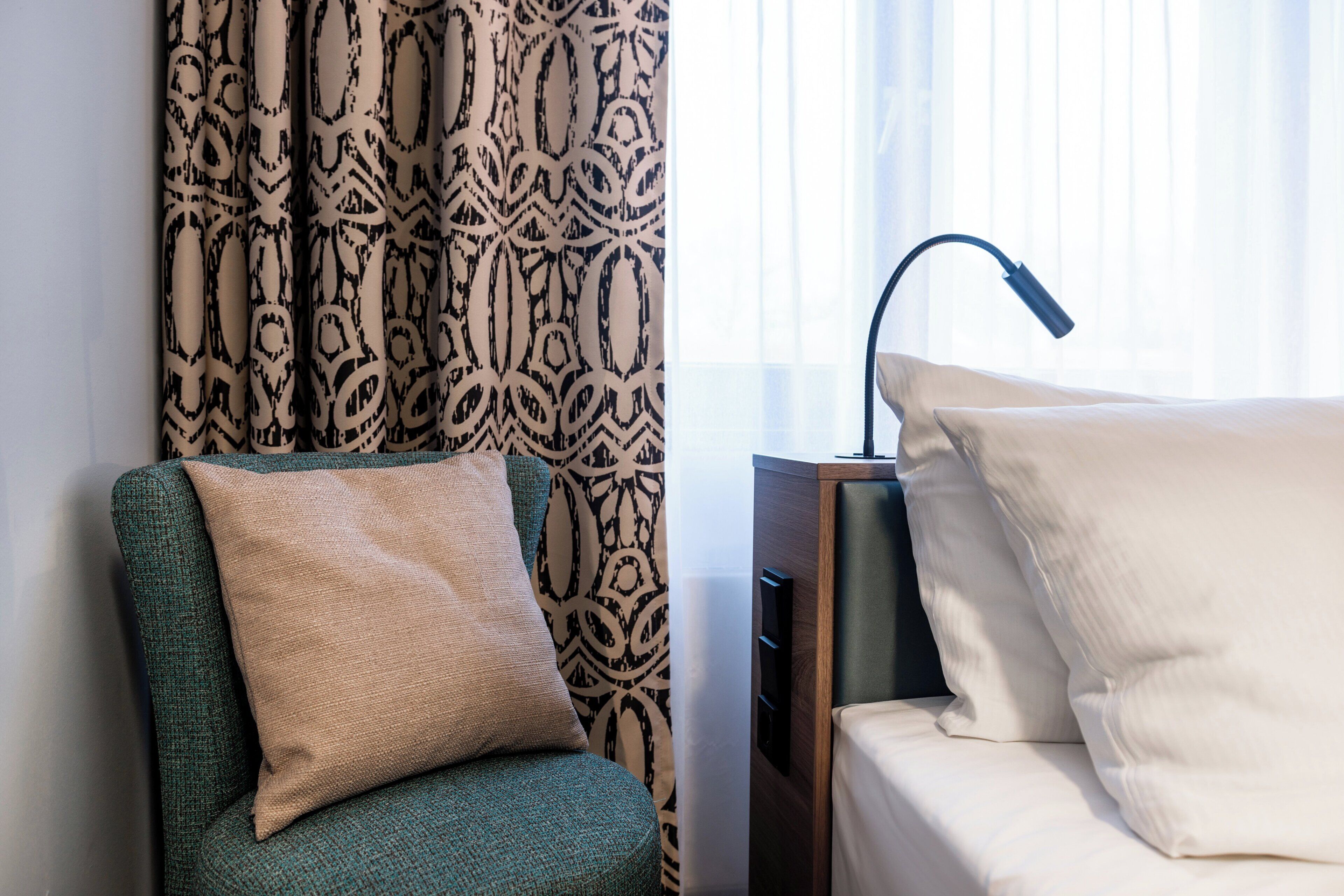 Una camera d'hotel con una poltrona verde, un cuscino beige, una tenda a motivi, una lampada da comodino e un letto con cuscini bianchi.