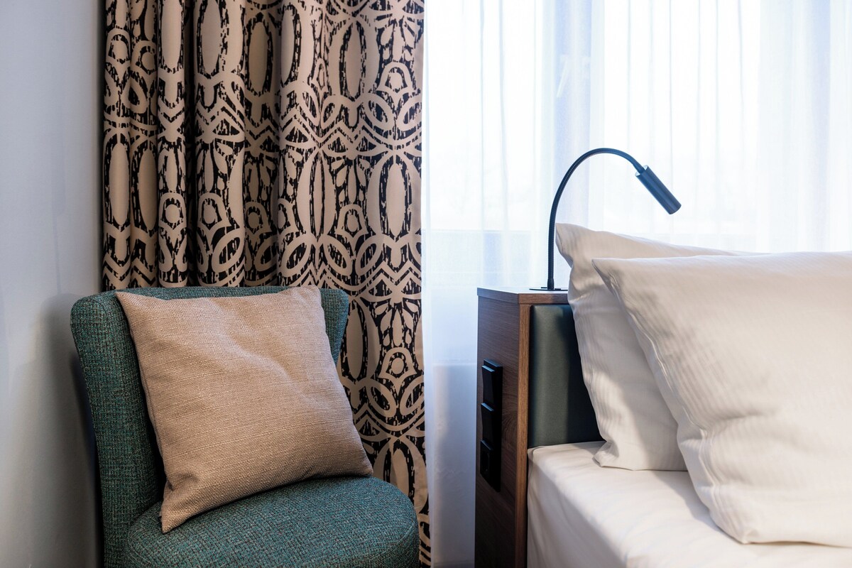 A hotel room with a green armchair, a beige pillow, a patterned curtain, a bedside lamp, and a bed with white pillows.