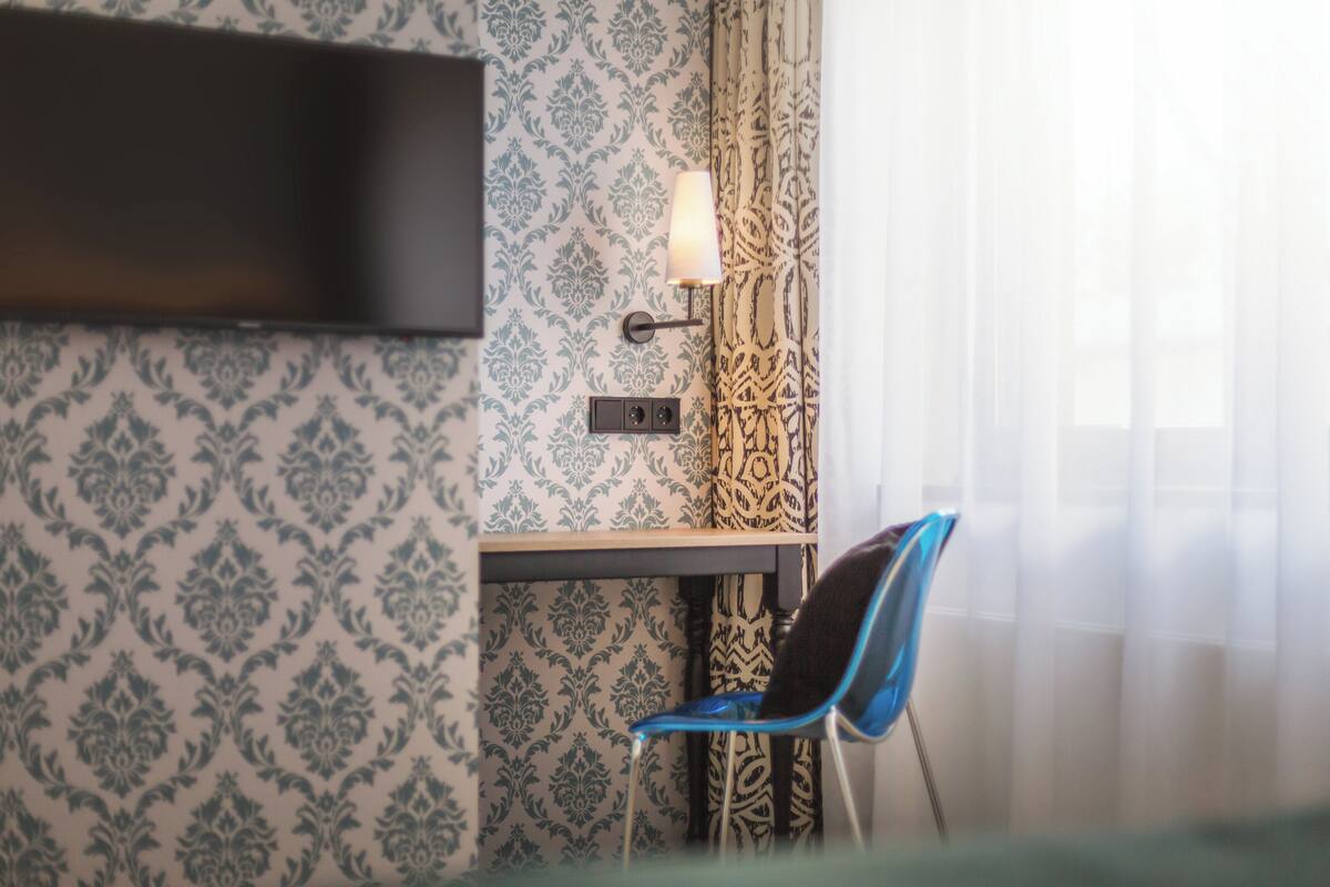 Hotellihuone, jossa on kuviollinen tapetti, seinälle asennettu taulutelevisio, työpöytä lampun kanssa ja sininen tuoli.