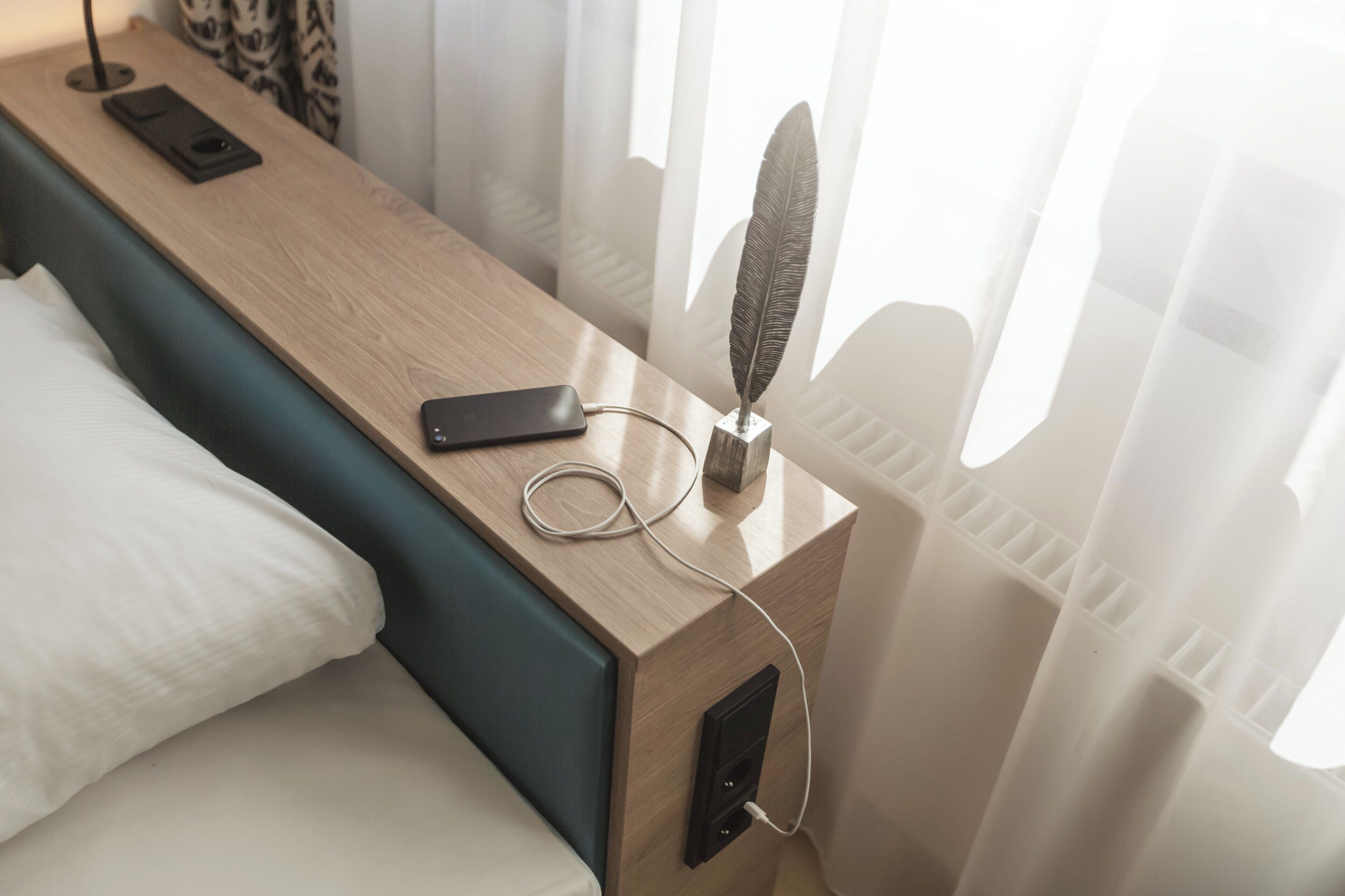 A wooden bedside table with a smartphone, charger, and decorative feather.