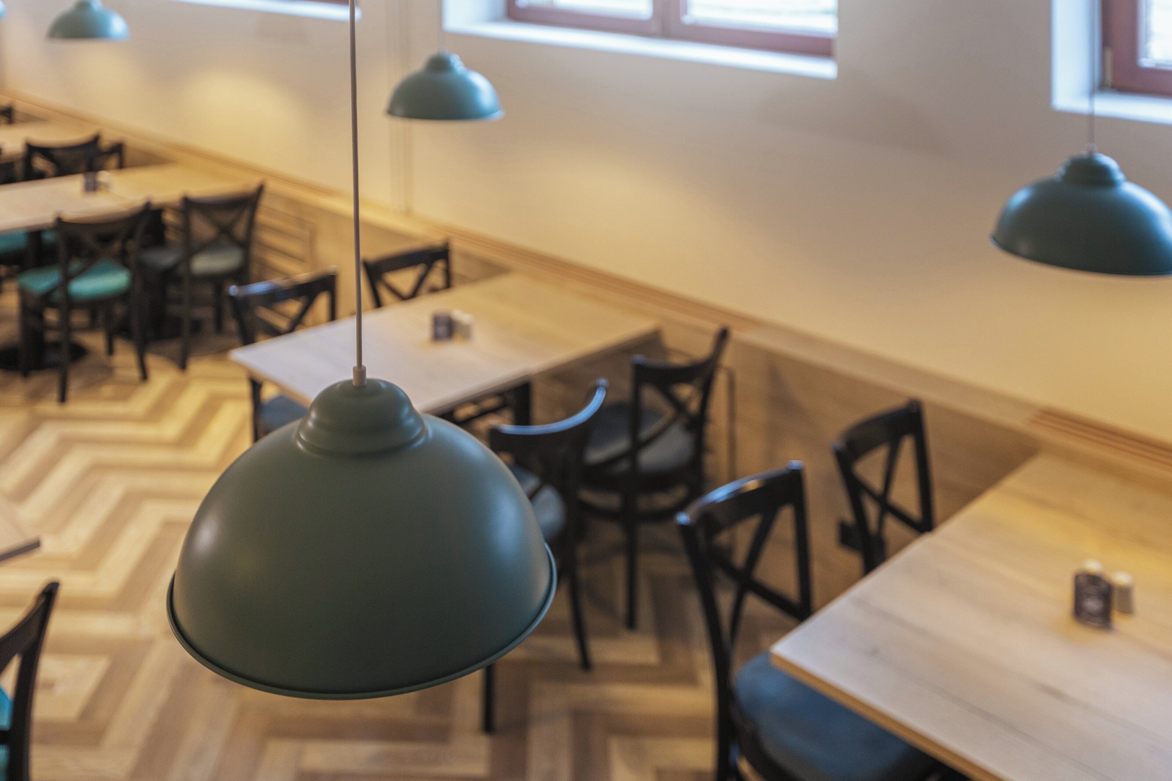 A restaurant interior with wooden tables and chairs, hanging green pendant lights, and a herringbone floor pattern.