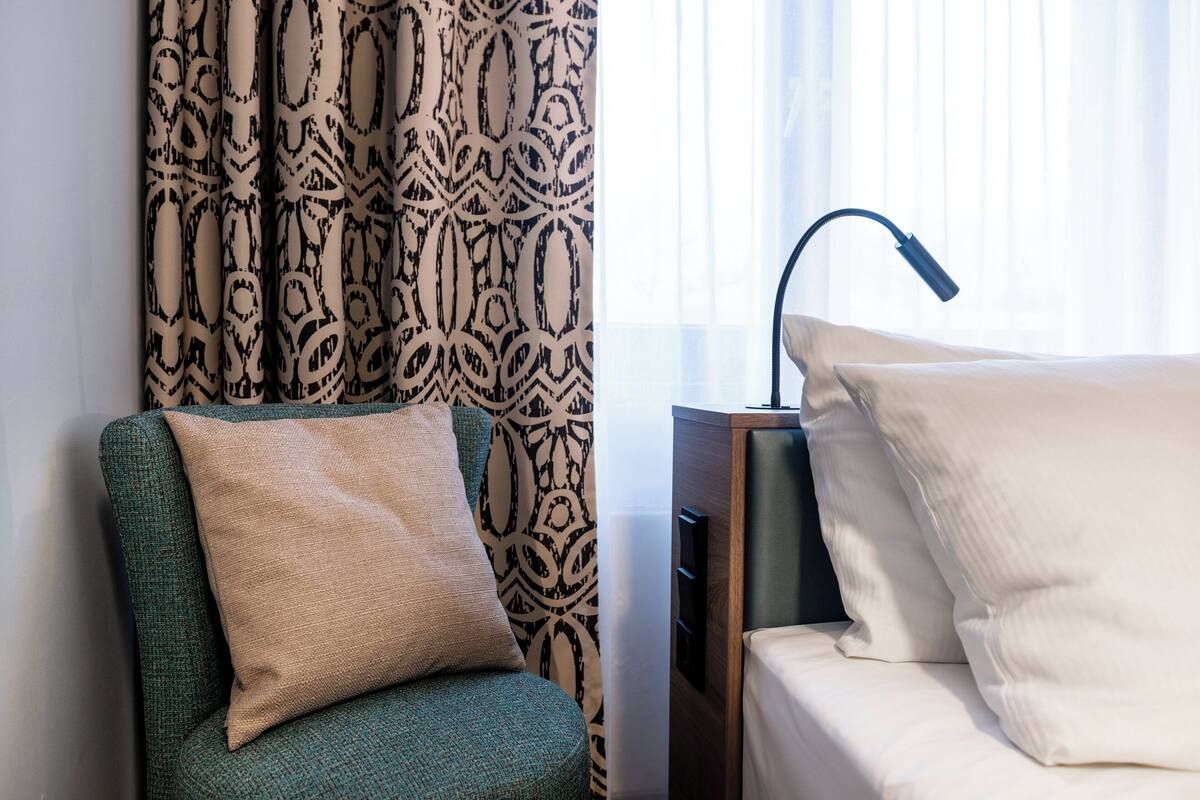 Hotellihuone, jossa on vihreä nojatuoli, beigenvärinen tyyny, kuviollinen verho, yöpöytälamppu ja valkoisilla tyynyillä varustettu sänky.